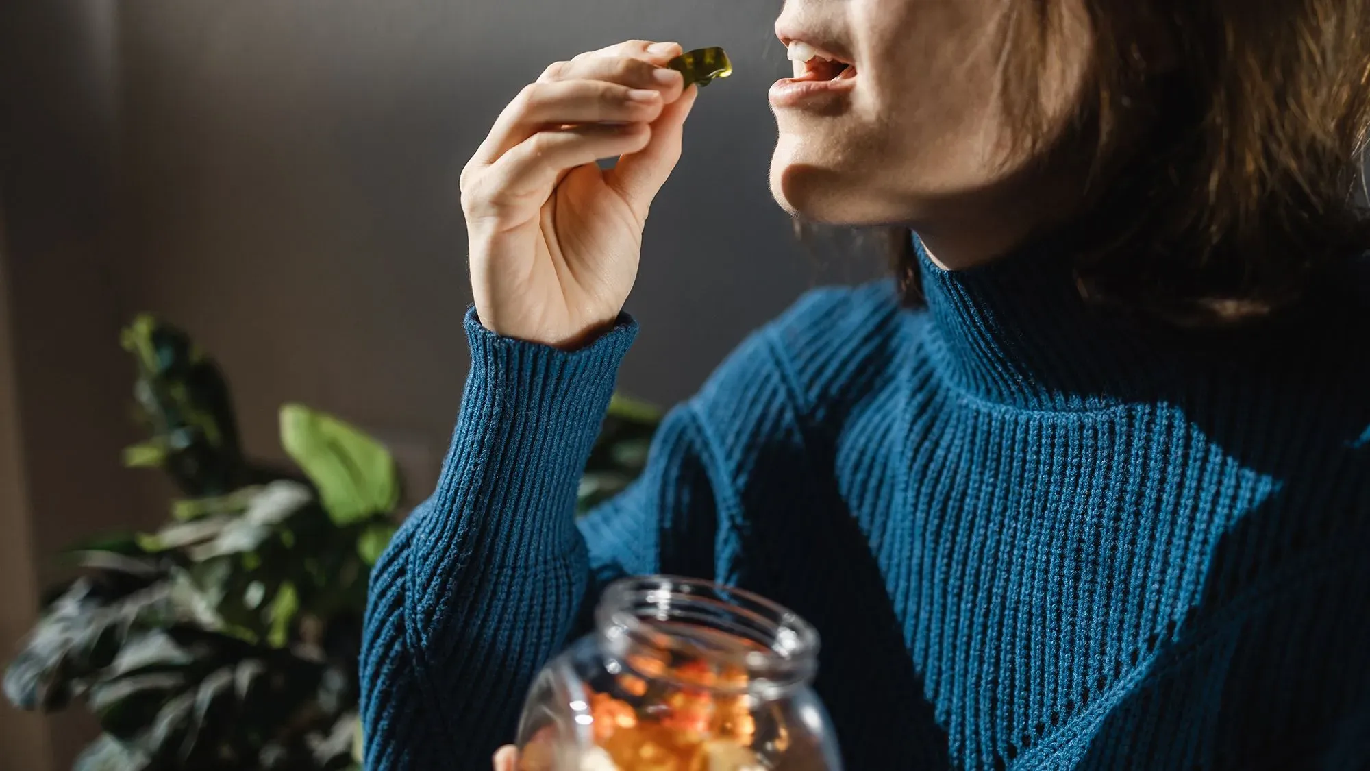 امرأة ترتدي سترة زرقاء، تتناول كبسولة من زجاجة تحتوي على مستخلصات القنب، مع وجود نباتات في الخلفية.