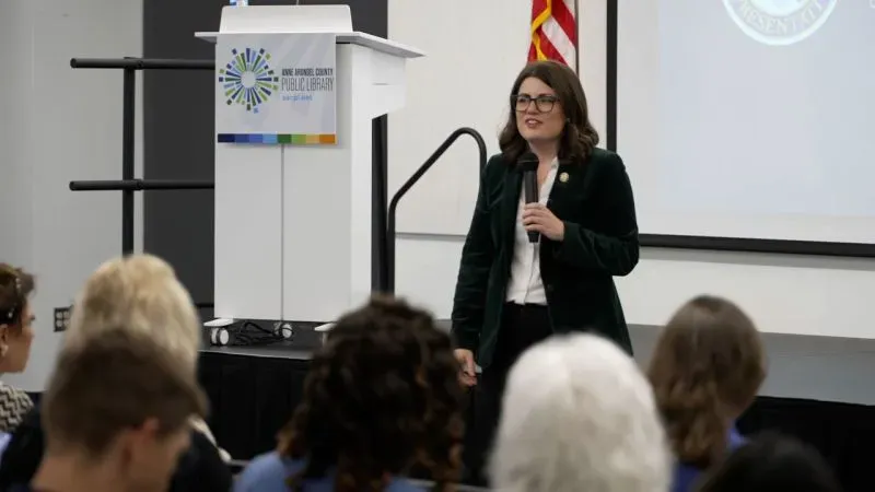 النائبة مارجوري تايلور غرين تتحدث في فعالية عامة، مع جمهور يستمع بتركيز. الخلفية تحتوي على شعار مكتبة عامة.