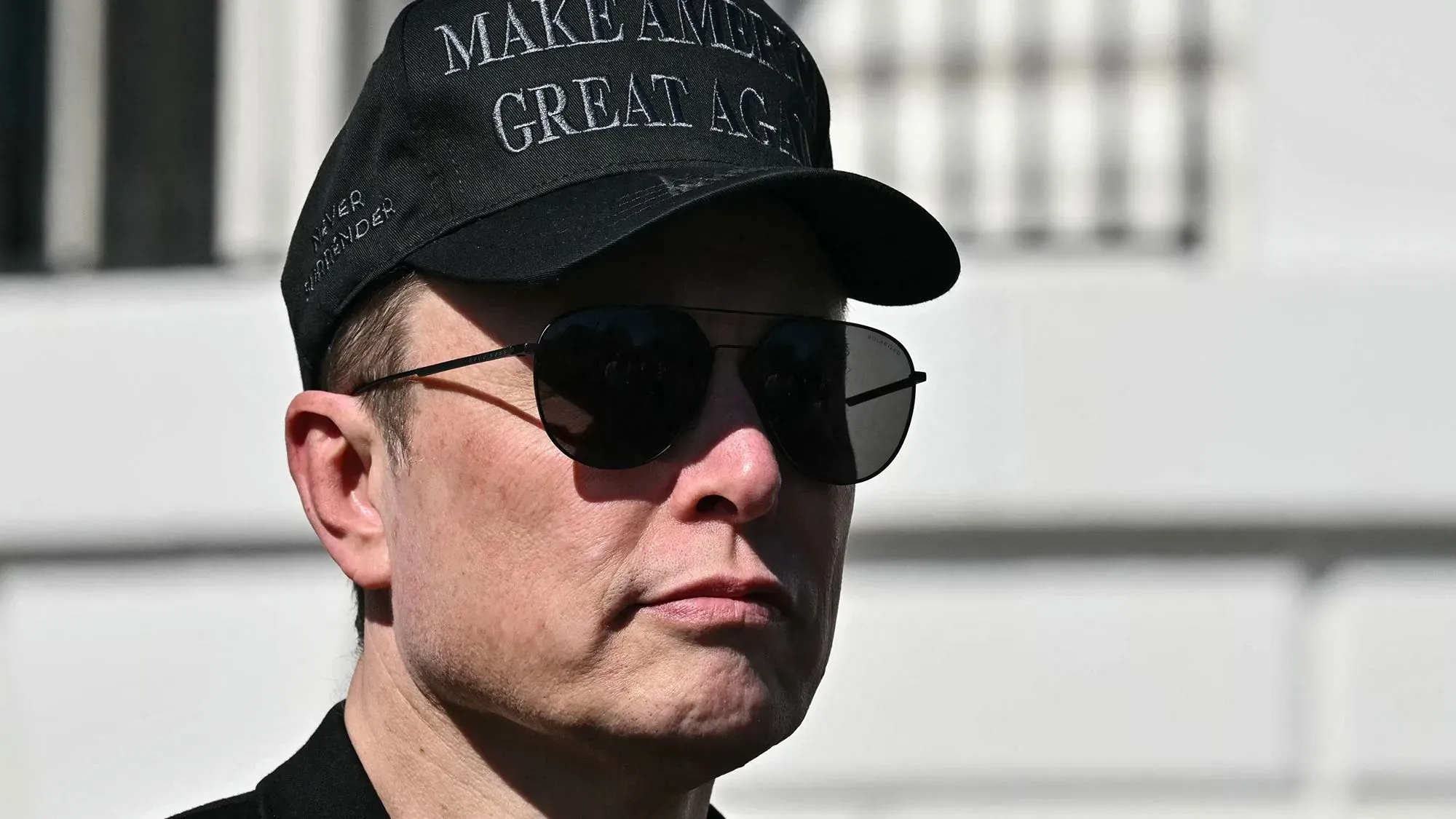 إيلون ماسك يرتدي نظارات شمسية وقبعة تحمل عبارة "اجعل أمريكا عظيمة مرة أخرى"، يعكس التوترات حول إدارة تسلا وأدائها.