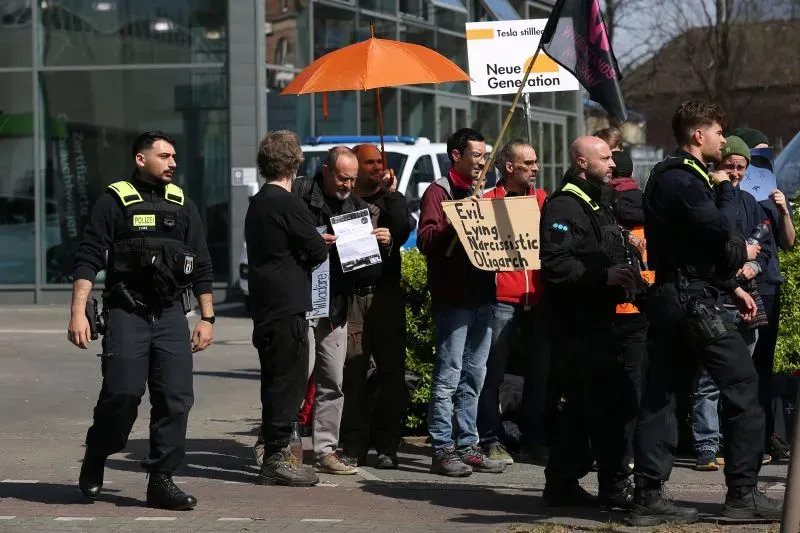 احتجاجات ضد تسلا في أوروبا، حيث يحمل المتظاهرون لافتات تعبر عن استيائهم من سياسة إيلون ماسك، مع وجود شرطة في الخلفية.