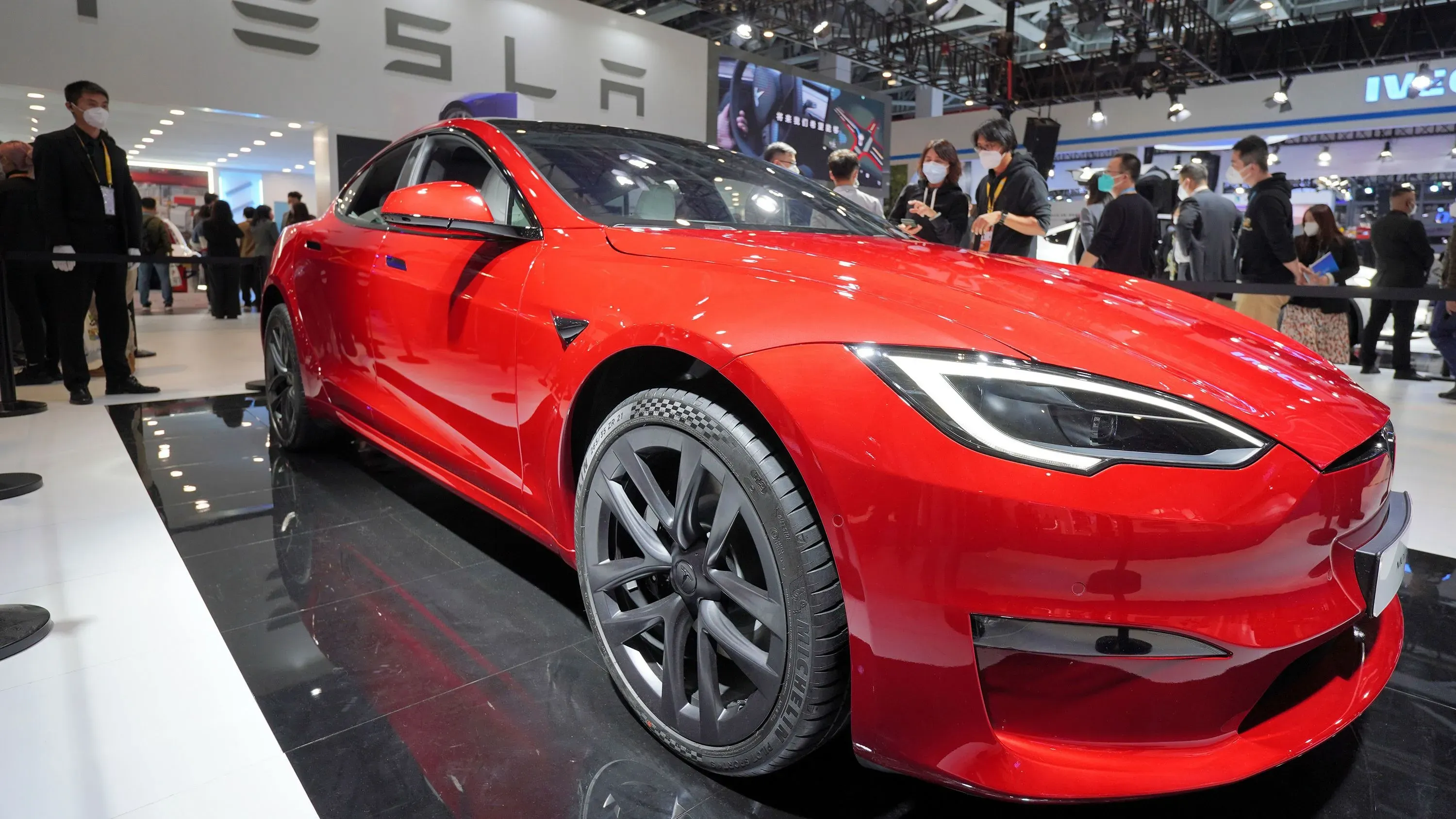 سيارة تسلا موديل S الحمراء تعرض في معرض سيارات، مع وجود زوار خلفها. تشير الصورة إلى تأثير الحرب التجارية على مبيعات السيارات الكهربائية.