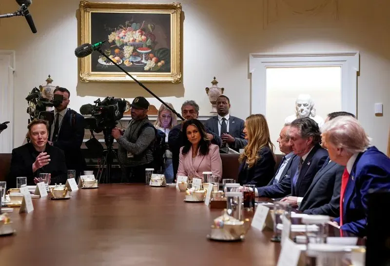 إيلون ماسك يتحدث خلال اجتماع مع مسؤولين في البيت الأبيض، محاطًا بالصحفيين، مما يعكس تأثير السياسة على تسلا.