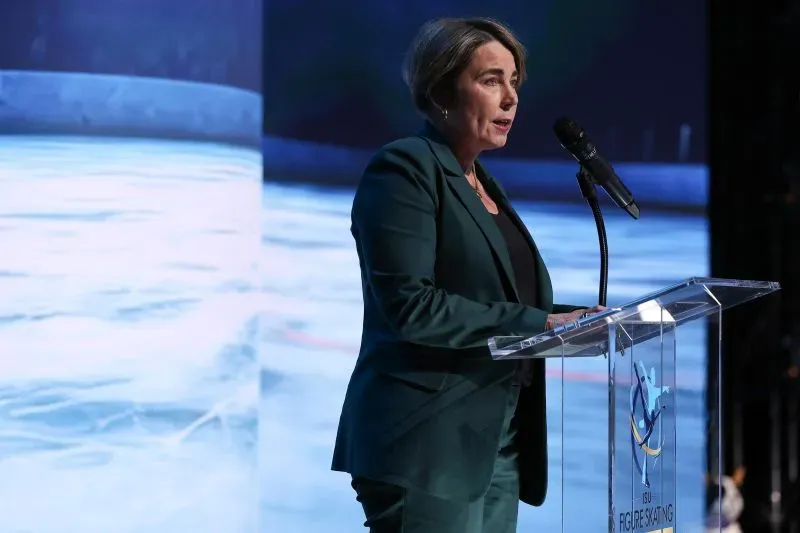 امرأة ترتدي بدلة خضراء تقف خلف ميكروفون على منصة، تتحدث خلال حدث متعلق بالتزلج الفني، مع خلفية مائية.