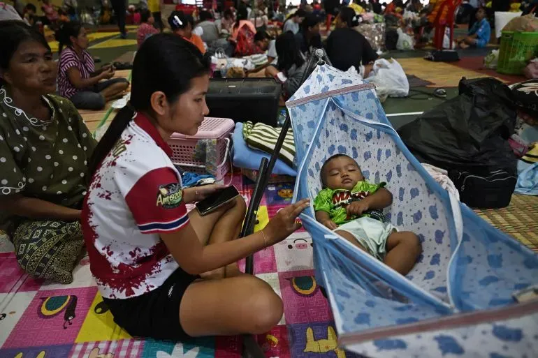 امرأة تجلس في مركز إيواء، تحمل هاتفها، بينما ينام طفلها في سرير صغير بجانبها. يظهر في الخلفية أشخاص آخرون في المكان.