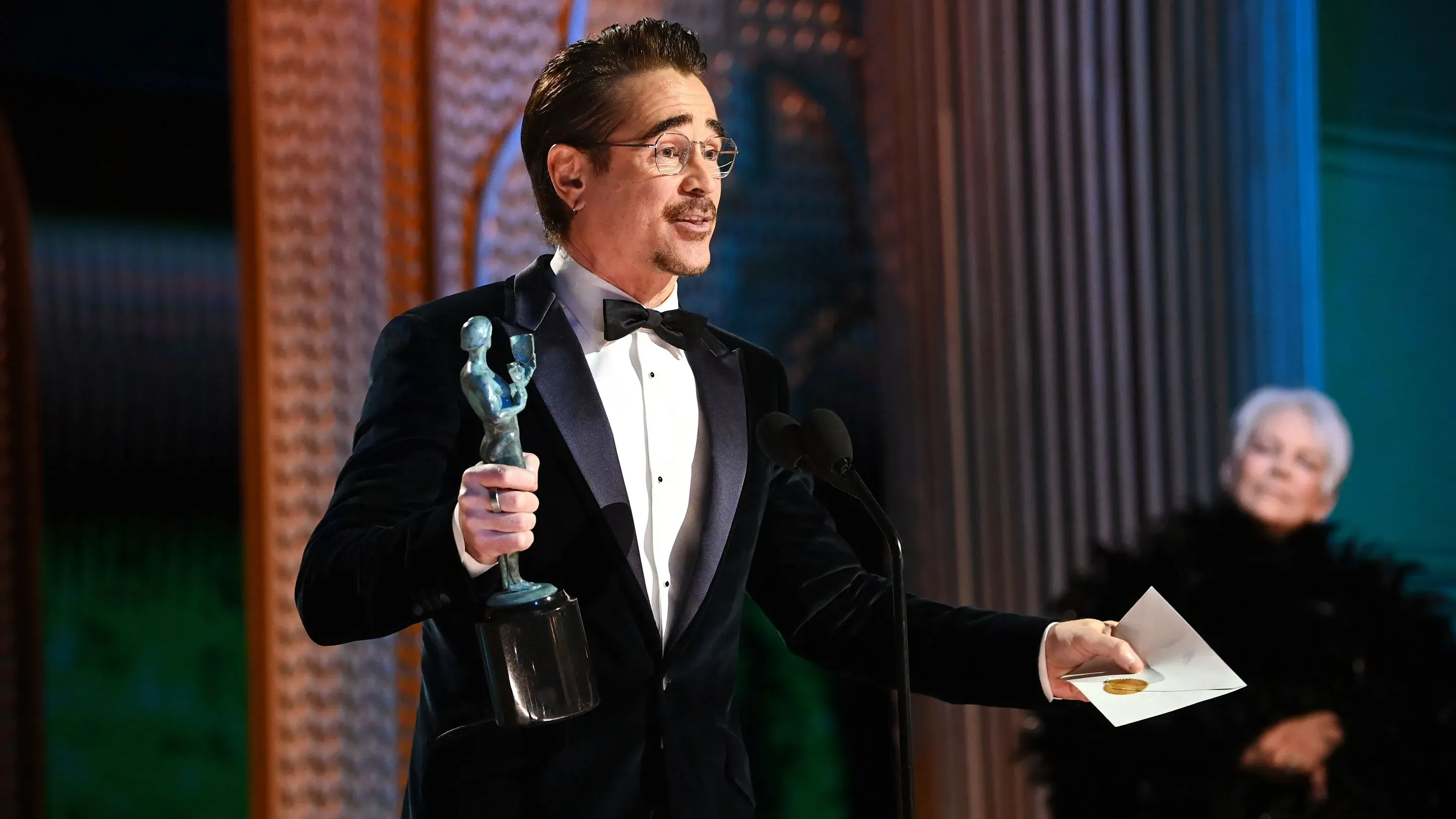 كولين فاريل يتسلم جائزة SAG، حاملاً تمثال الممثل، بينما تظهر خلفه جودي دينش. الحفل يعكس لحظات من الكوميديا والجدية.