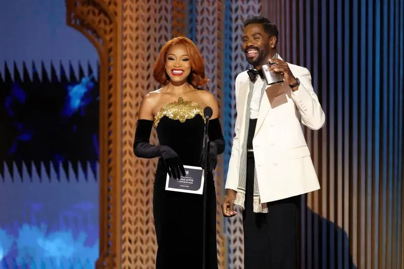 مقدمة حفل توزيع جوائز SAG، ترتدي فستانًا أسود مع تفاصيل ذهبية، تتحدث مع زميلها الذي يحمل جائزة، وسط أجواء احتفالية.