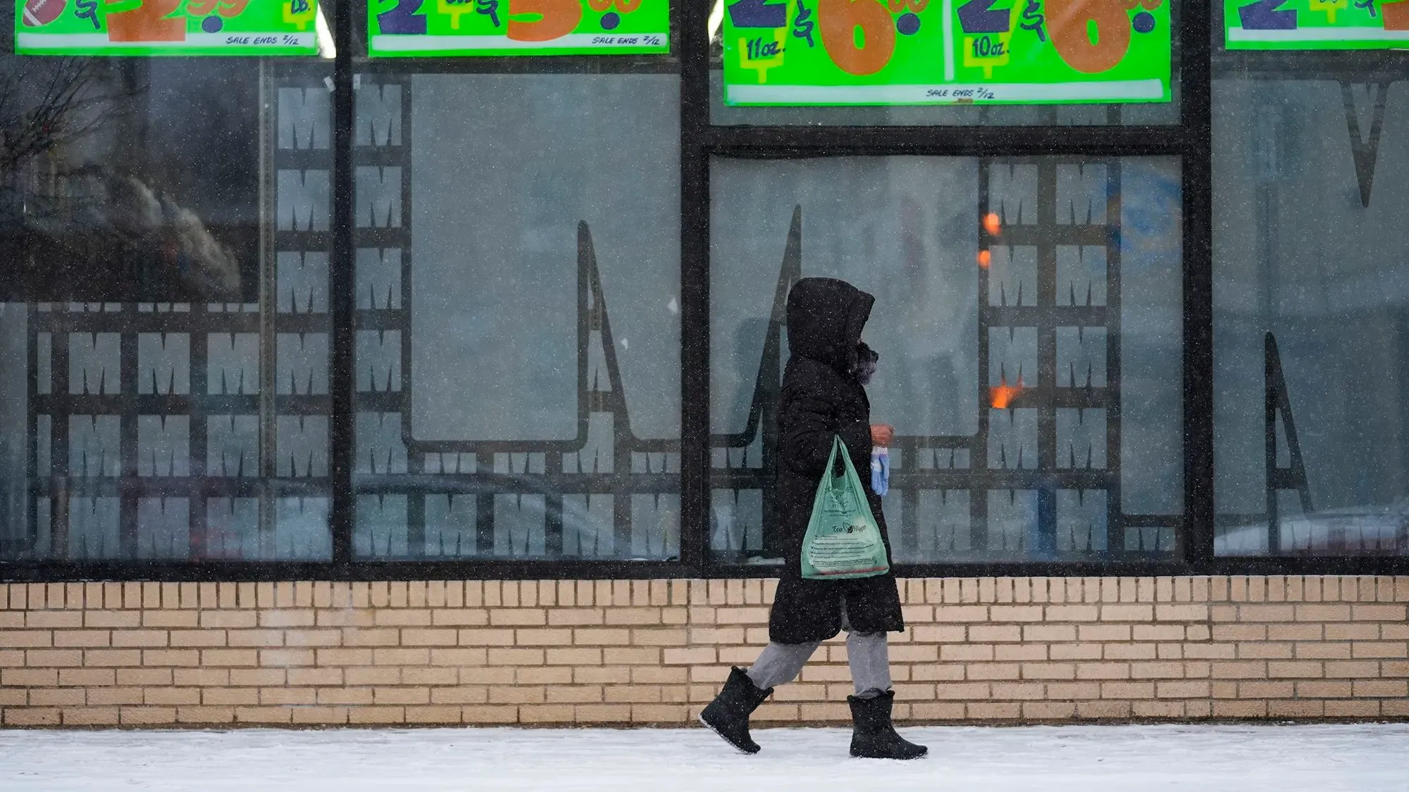 امرأة ترتدي معطفًا أسود وتمشي في الثلوج بجوار واجهة متجر، حيث تظهر علامات تخفيضات ملونة تعكس تراجع الثقة الاقتصادية.