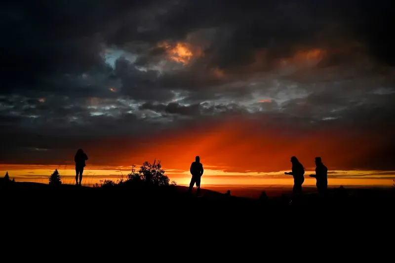 ظلال أشخاص يقفون على قمة تلة، يستمتعون بغروب الشمس المذهل خلف سحب داكنة، مما يعكس حالة عدم اليقين في السفر.