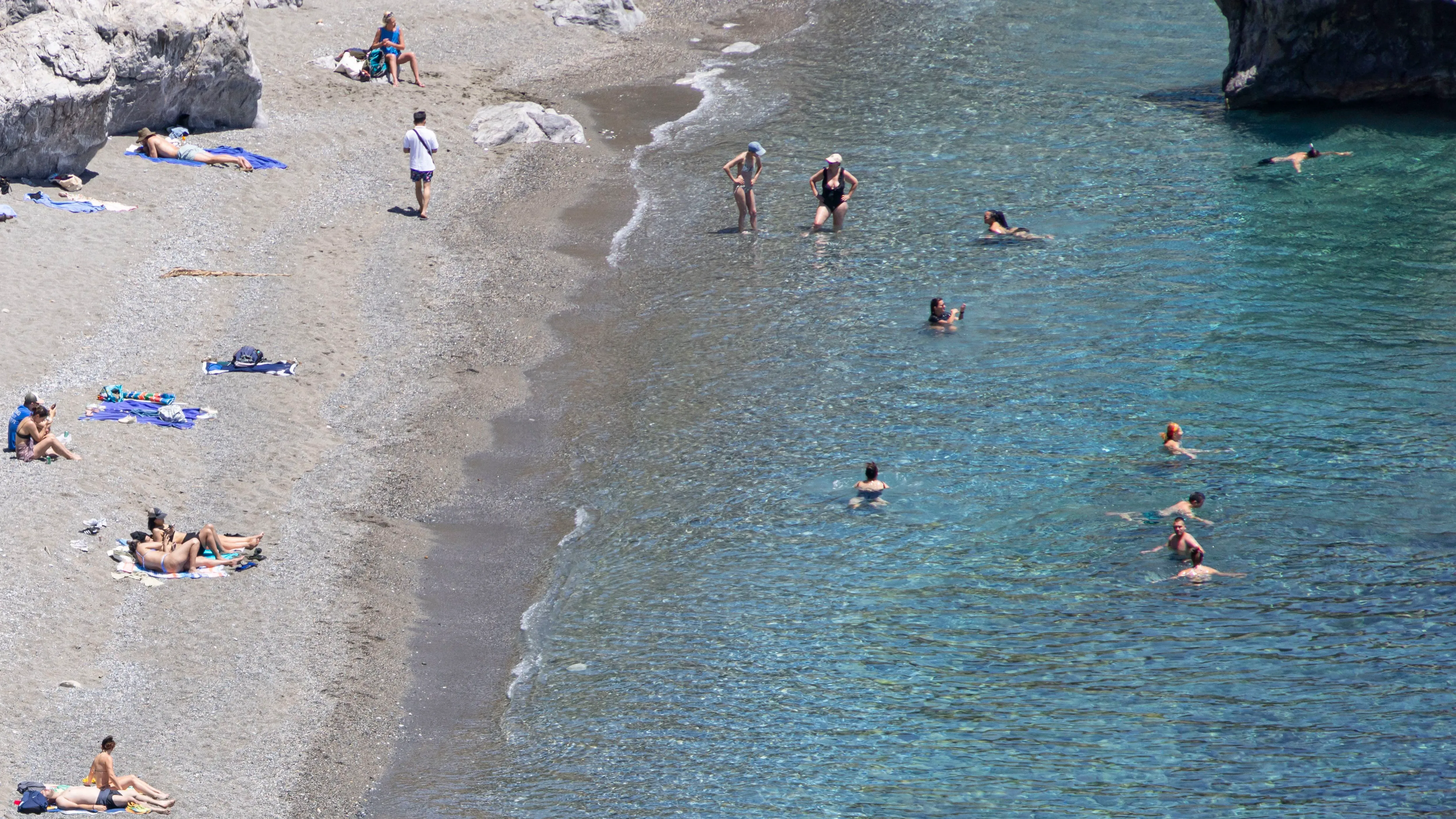 شاطئ يضم مجموعة من الأشخاص يستمتعون بالشمس والماء، مع وجود بعضهم على الرمال وآخرين يسبحون في البحر.