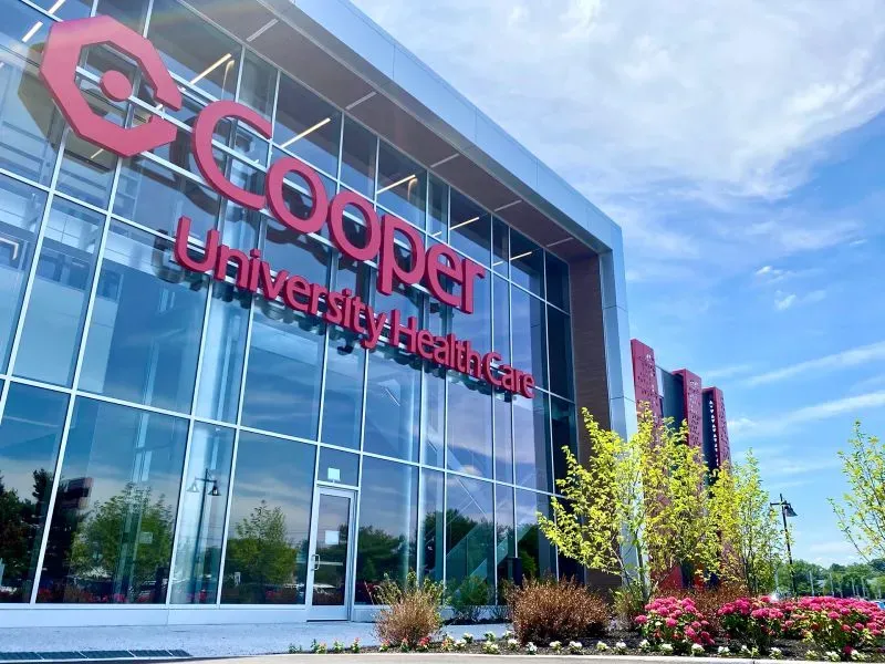 واجهة مركز Cooper University Health Care مع لافتة كبيرة، محاطة بأشجار وزهور، تعكس تجديد مراكز التسوق لتشمل خدمات صحية.
