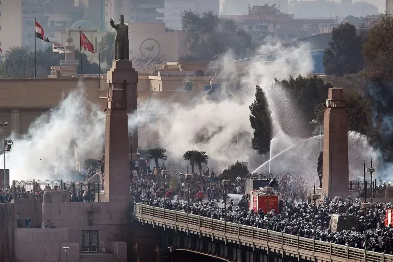 تجمع حشود كبيرة من المتظاهرين في وسط القاهرة، وسط دخان الغاز المسيل للدموع، مع وجود تمثال بارز في الخلفية.
