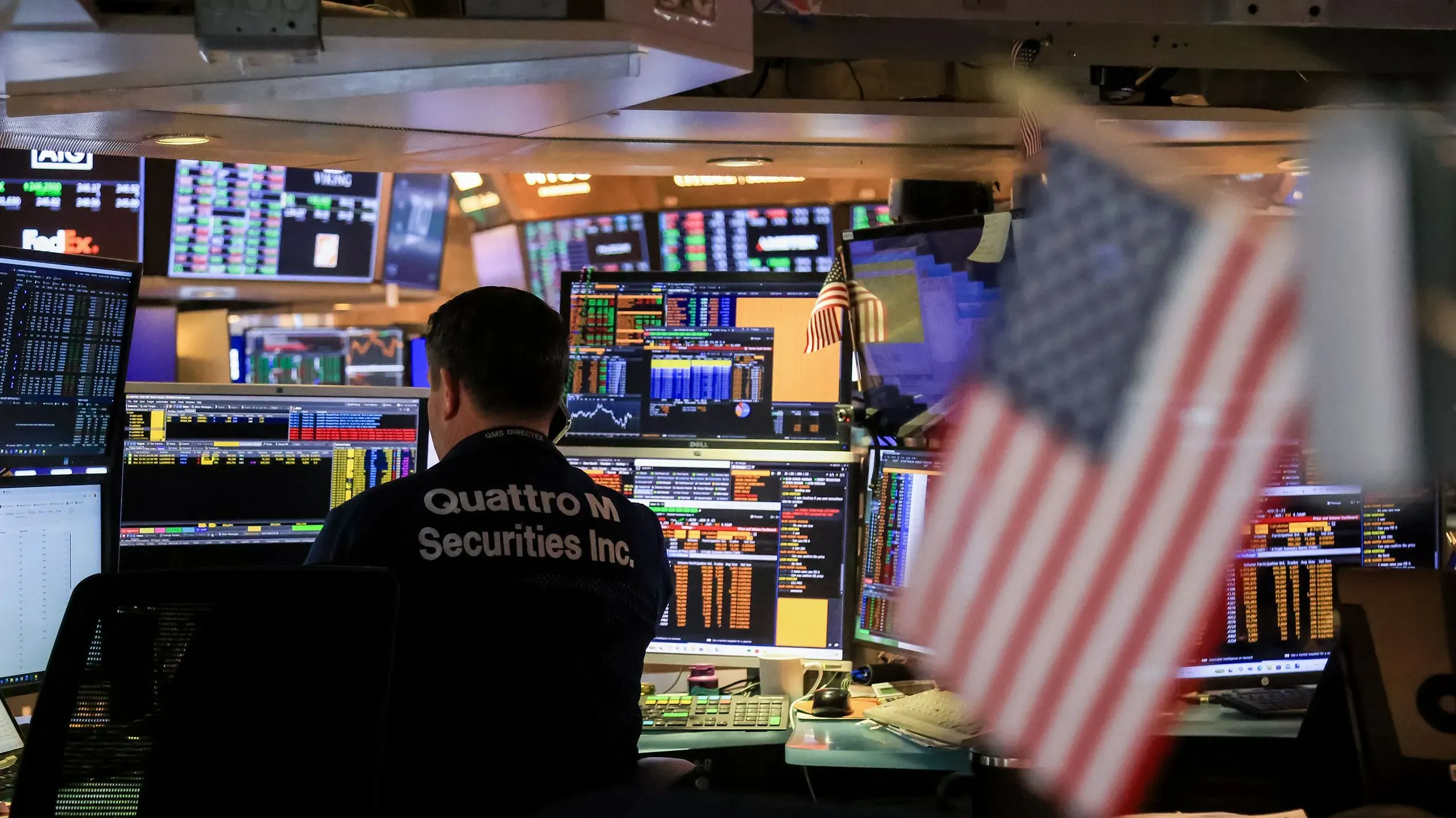 متداول في مركز مالي، محاط بشاشات تعرض بيانات السوق وأسعار الأسهم، مع وجود علم أمريكي في الخلفية.