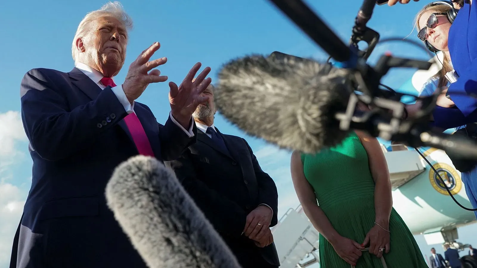 دونالد ترامب يتحدث للصحفيين أمام طائرة، محاطًا بكاميرات وميكروفونات، وسط أجواء مشحونة بعد إقالة مورين كومي.