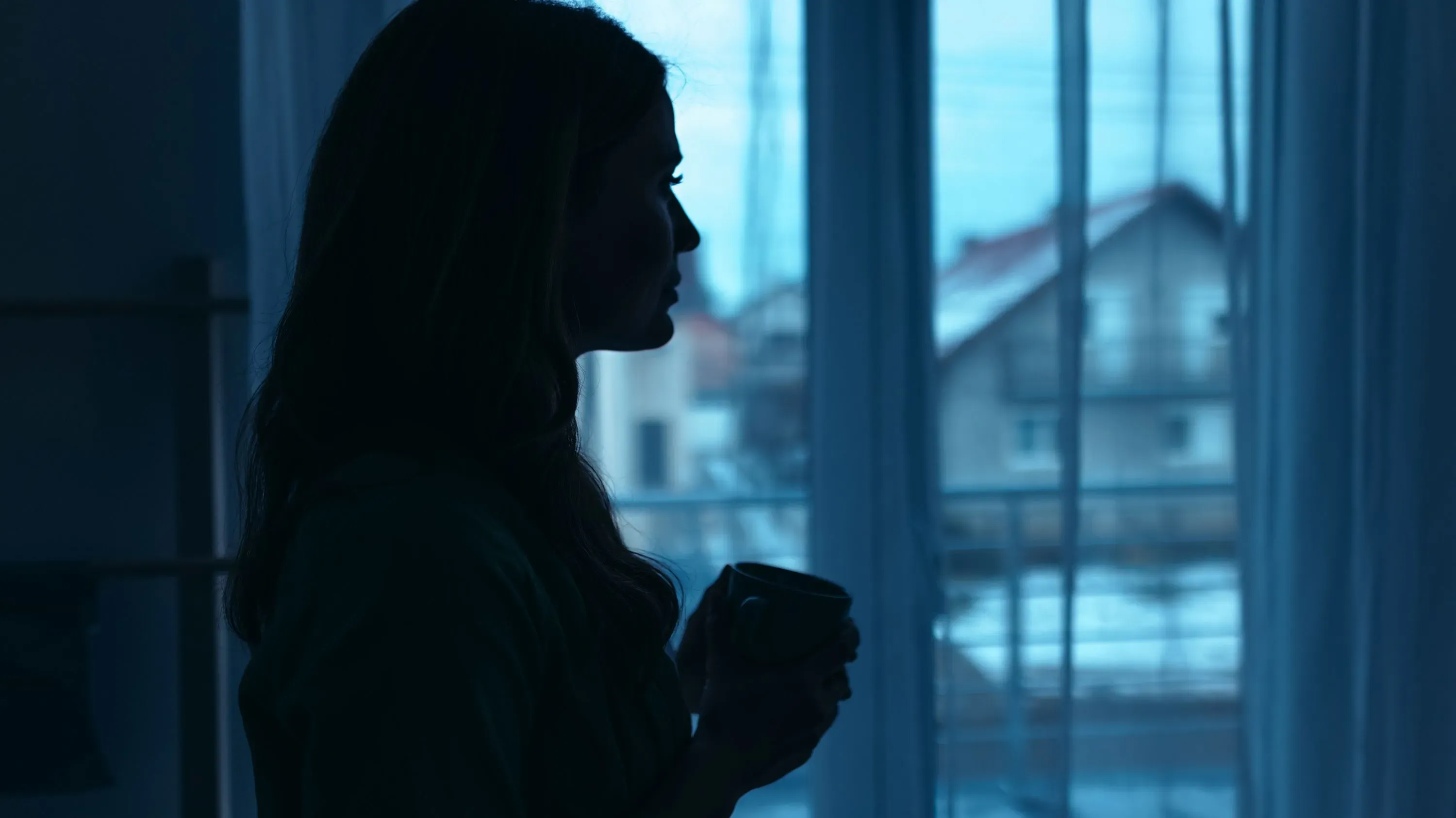 امرأة واقفة بجانب نافذة، تحمل كوبًا، وتبدو silhouetted في ظلال زرقاء، تعكس شعور الكآبة المرتبطة بيوم الاثنين الأزرق.