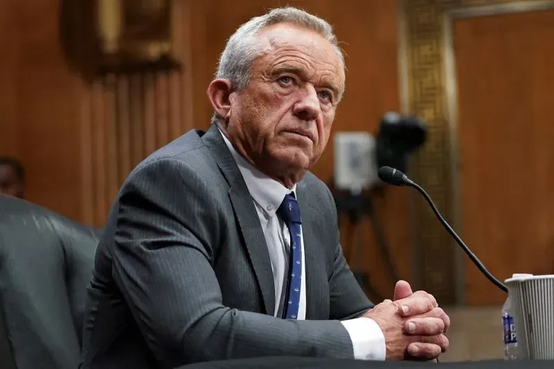 رجل مسن يجلس في قاعة جلسات، يبدو عليه التفكير العميق، يرتدي بدلة رسمية، مع وجود ميكروفون أمامه، في سياق مناقشة حول اللقاحات.