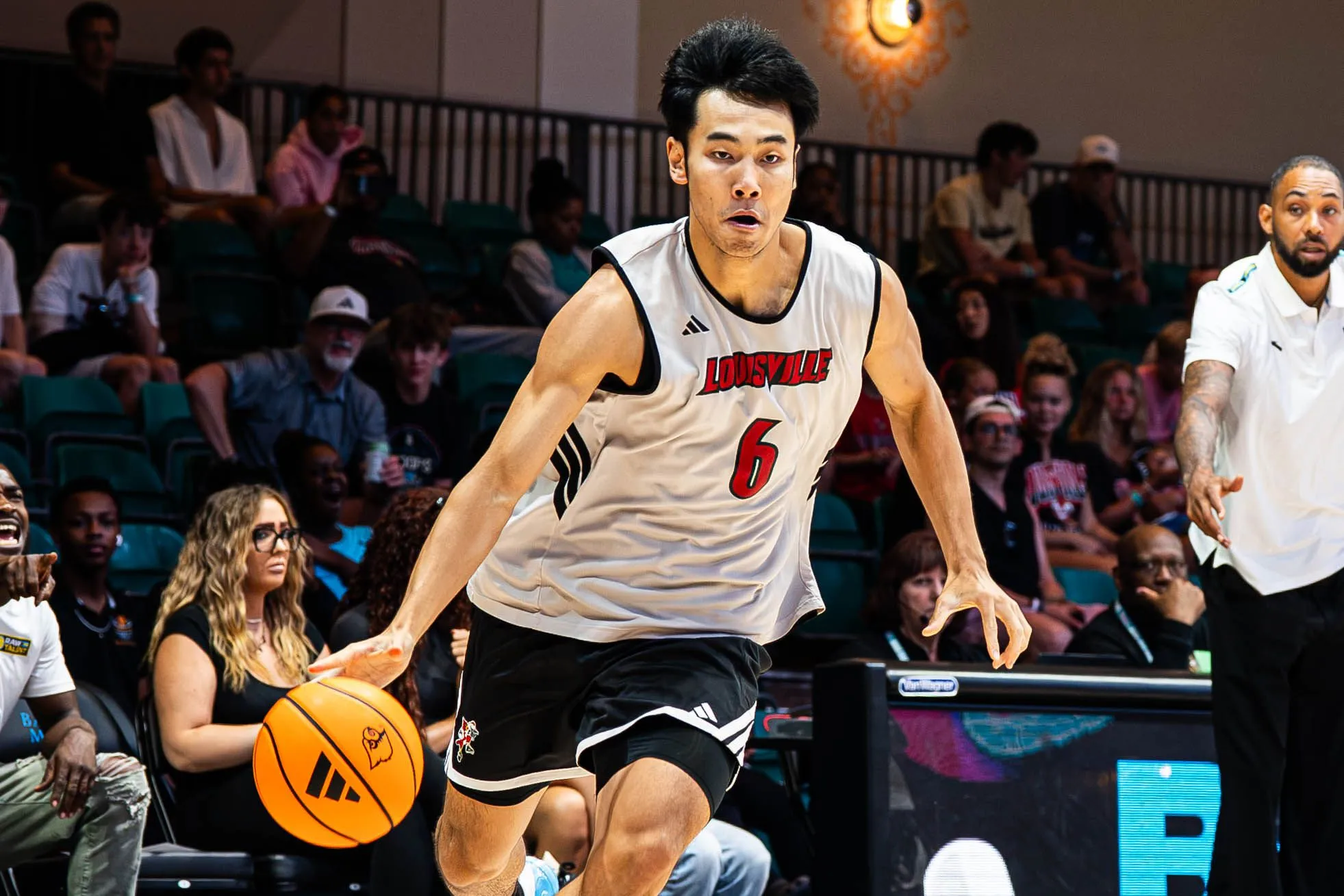 لاعب كرة السلة الصيني Zhang Boyuan يرتدي قميص Louisville Cardinals ويحمل الكرة خلال مباراة في NCAA، مع جمهور ومشجعين في الخلفية.