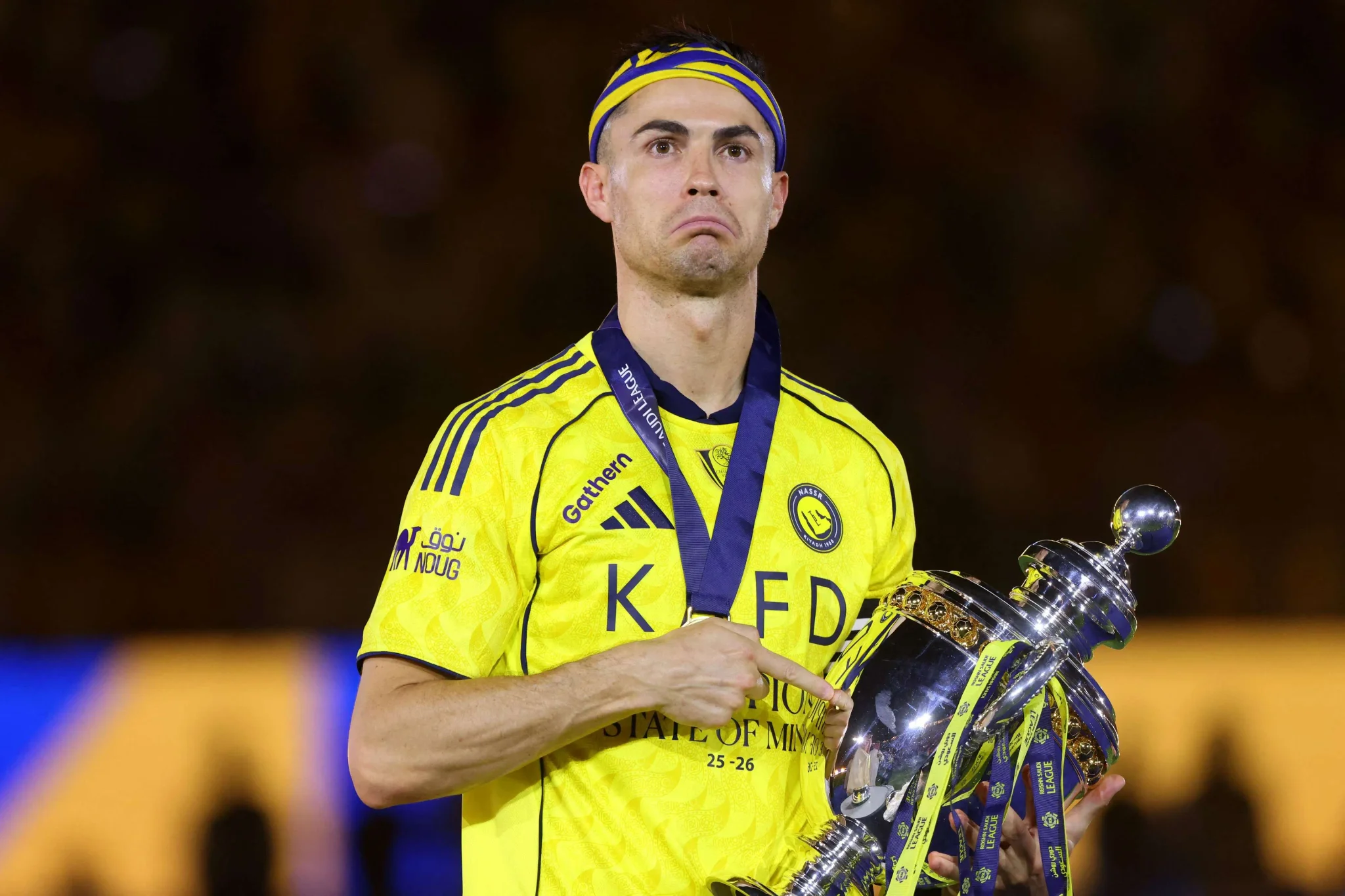 لاعب كرة قدم يرتدي زي أصفر يحمل كأس البطولة مع ربطة رأس وشريط ميدالية، يعبر عن فخره وانتصاره في موسم 2026-2027.