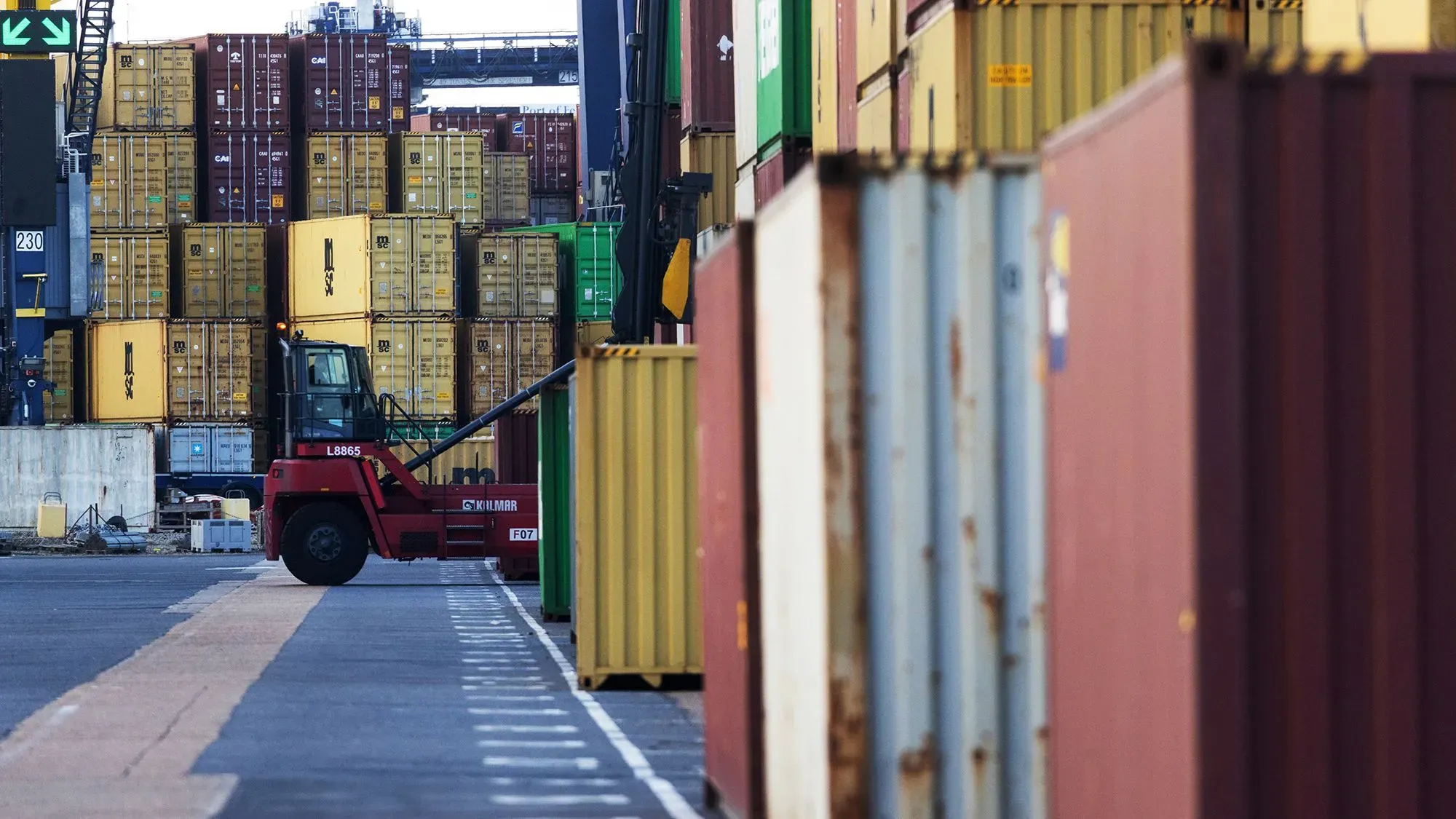 تظهر الصورة شاحنة نقل تعمل في ميناء، محاطة بالحاويات الملونة، مما يعكس تأثير الرسوم الجمركية على التجارة الدولية.