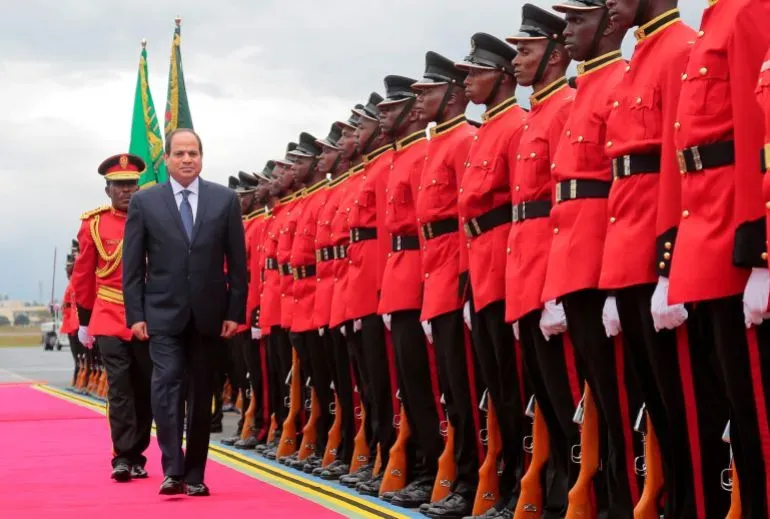 رجل يرتدي بدلة رسمية يسير على سجادة حمراء، محاطًا بمجموعة من الجنود في زي رسمي أحمر، خلال مراسم استقبال في تنزانيا.