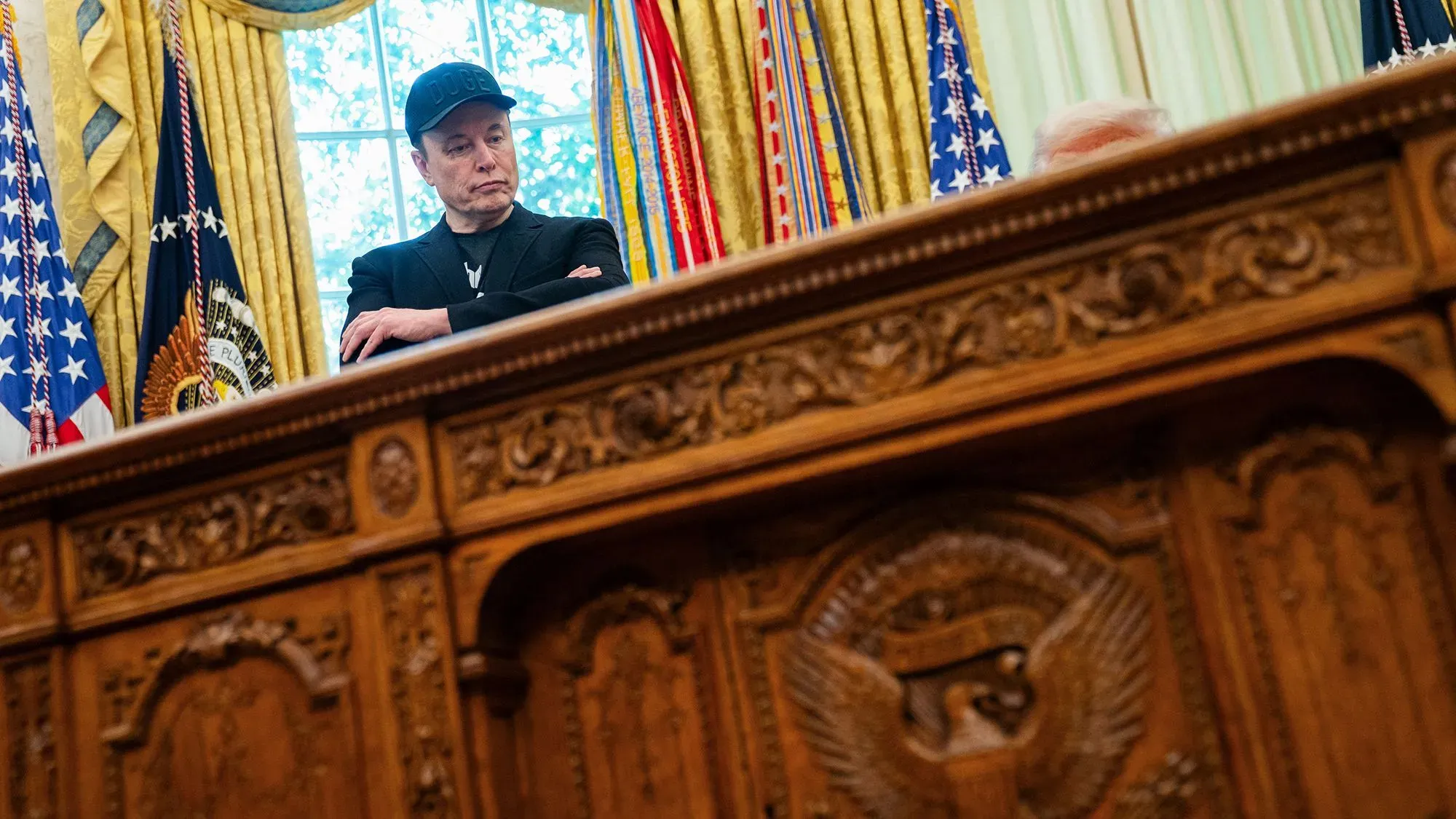 إيلون ماسك يقف في المكتب البيضاوي، معبرًا عن مشاعر التوتر أثناء مناقشة قضايا سياسية مع ترامب، في خلفية تحمل الأعلام الأمريكية.