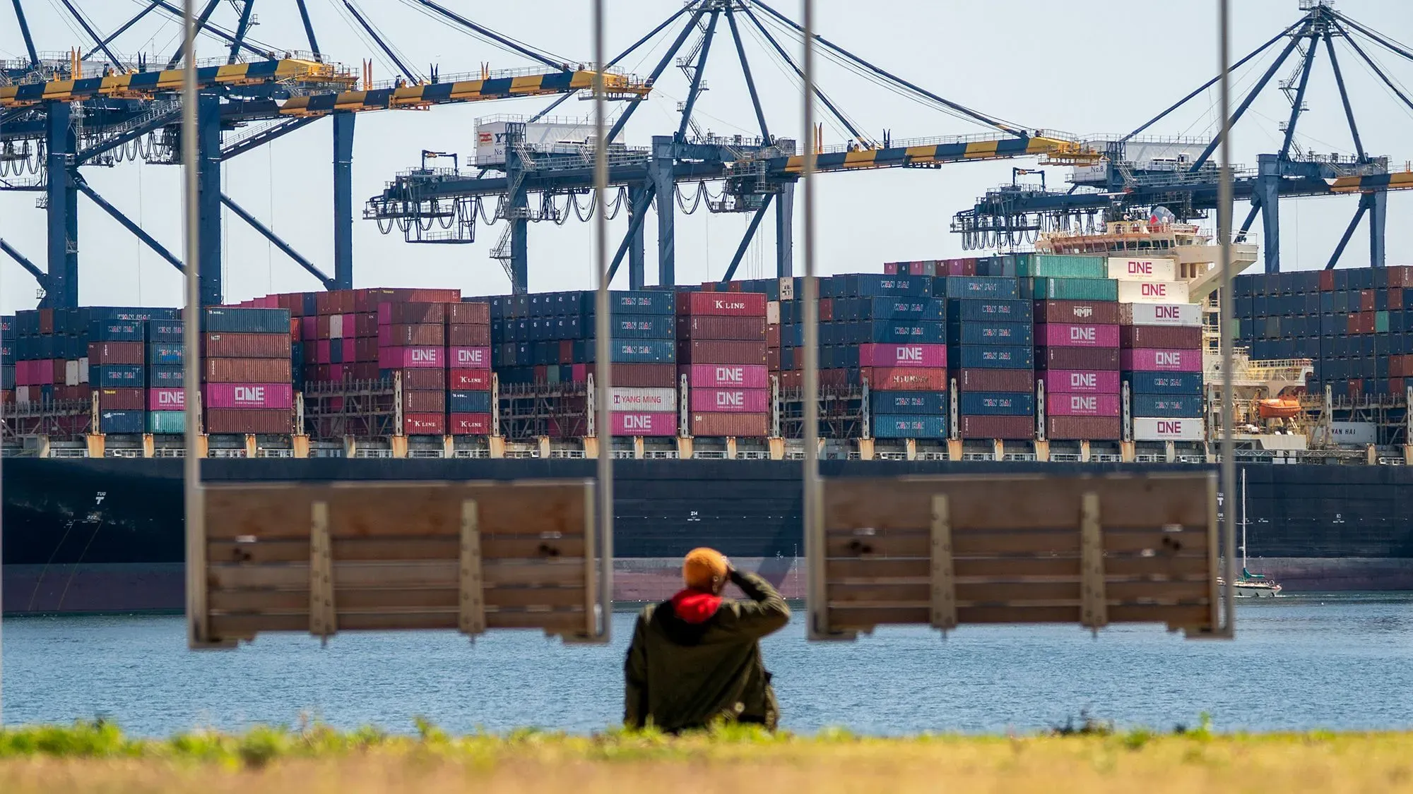 رجل يجلس على أرجوحة أمام ميناء مزدحم، حيث تظهر سفينة شحن محملة بالحاويات، مما يعكس تأثير الحرب التجارية على الأسواق.