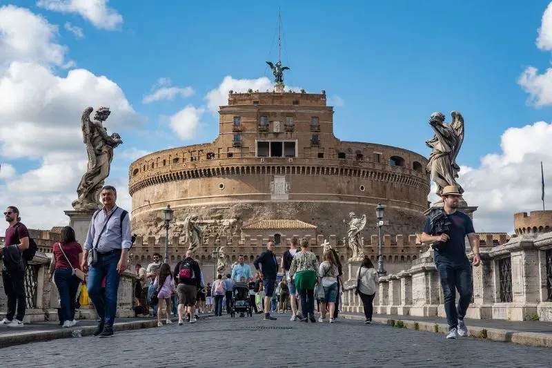 مجموعة من السياح يسيرون بجوار قلعة سانت أنجلو في روما، تحت سماء زرقاء مع سحب بيضاء، مما يعكس أجواء الصيف النابضة بالحياة.