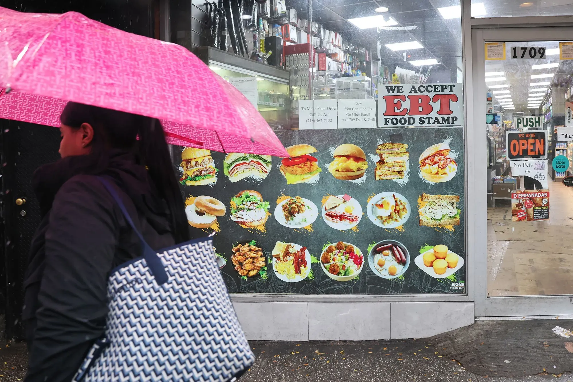 امرأة تحمل مظلة وردية تسير بجوار واجهة متجر تعرض لافتة "نقبل بطاقات الطعام EBT" وصور أطباق متنوعة، تعبيرًا عن تأثير الإغلاق الحكومي على الإعانات الغذائية.