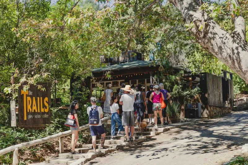 طابور من الزوار أمام كافيتيريا "ذا تريلز" في حديقة غريفيث بارك، محاط بأشجار خضراء، حيث يستمتعون بأجواء الطبيعة.