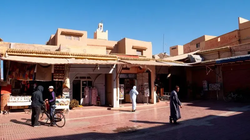 سوق تقليدي في تارودانت، المغرب، يظهر محلات بيع الحرف اليدوية والتوابل، مع سكان يتجولون في الأزقة تحت سماء زرقاء.