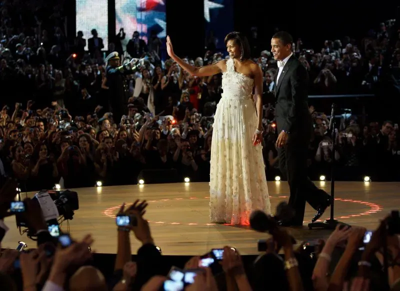 ميشيل أوباما ترتدي فستانًا أبيض مزين بالورود والكريستال خلال حفل تنصيب باراك أوباما، محاطة بجمهور يلتقط الصور.