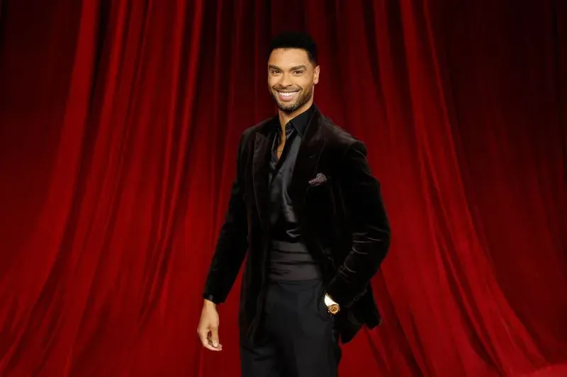 صورة لرجل يرتدي بدلة أنيقة، مبتسمًا أمام خلفية حمراء، مما يعكس جاذبية شخصيته المحتملة في دور "جيمس بوند".