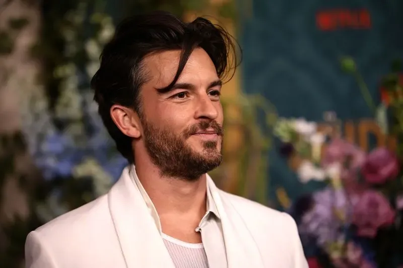 مظهر جذاب لممثل وسيم ذو لحية خفيفة وشعر مموج، يرتدي سترة بيضاء في حدث رسمي، مع خلفية مزهرة ملونة.