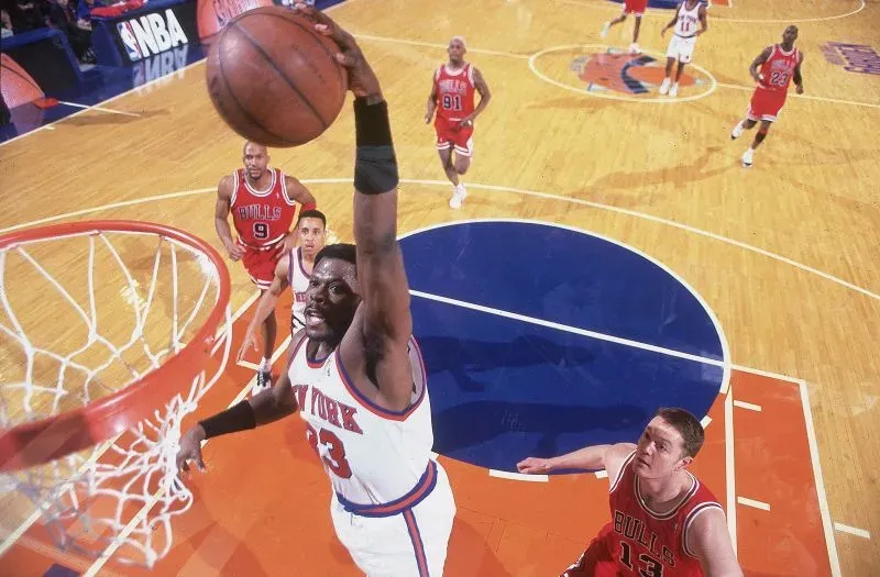 باتريك إيوينج يسجل سلة رائعة لفريق نيويورك نيكس خلال مباراة في التسعينيات، مع لاعبين من فريق شيكاغو بولز في الخلفية.