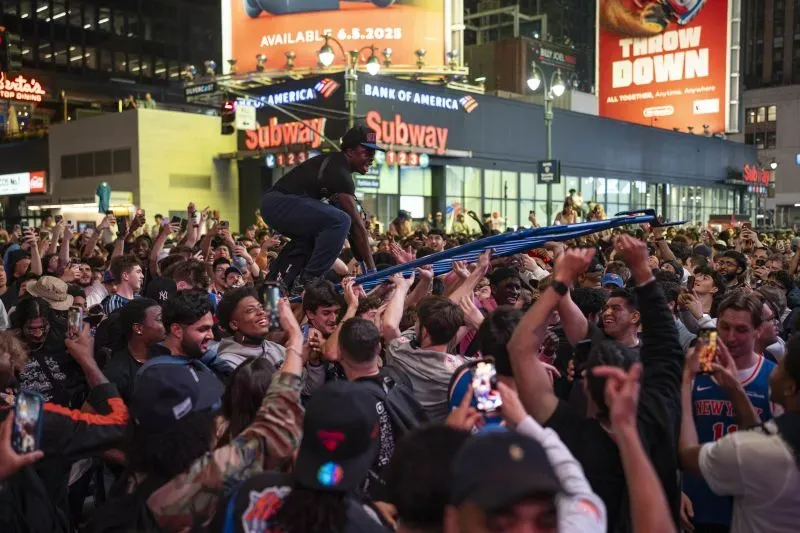 حشود من مشجعي نيويورك نيكس يحتفلون في الشارع بعد مباراة، مع شخص يتسلق عمودًا، مع أجواء حماسية تعكس شغفهم بالفريق.