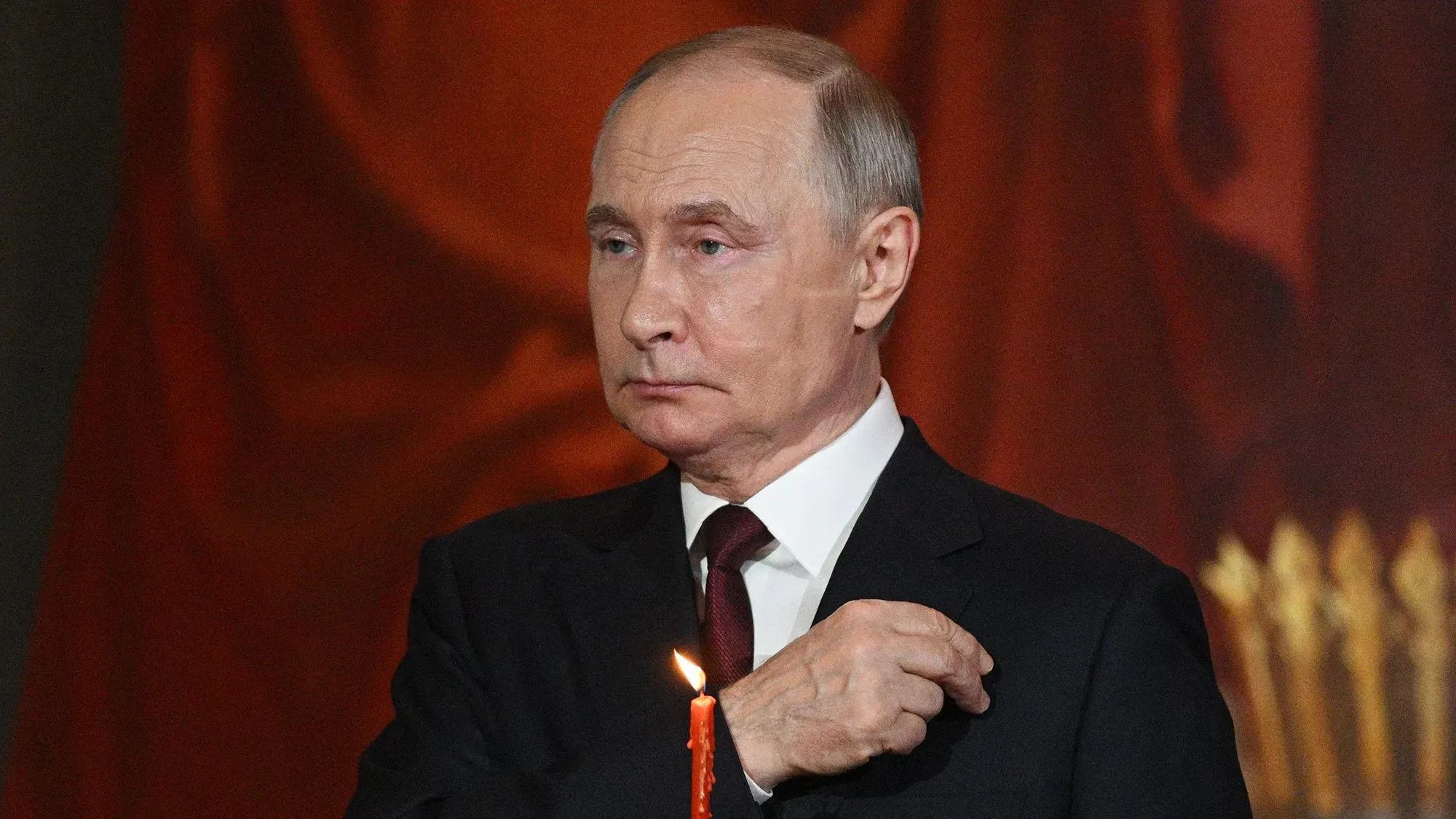 فلاديمير بوتين، الرئيس الروسي، يقف بجانب شمعة مضاءة، مع تعبير جاد، في خلفية حمراء، خلال حديث عن وقف إطلاق النار في أوكرانيا.