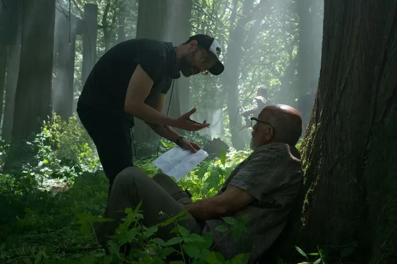 مشهد من كواليس "The Last of Us" يظهر مخرجًا يتحدث مع ممثل في غابة مضاءة بأشعة الشمس، مع تفاصيل طبيعية حولهم.