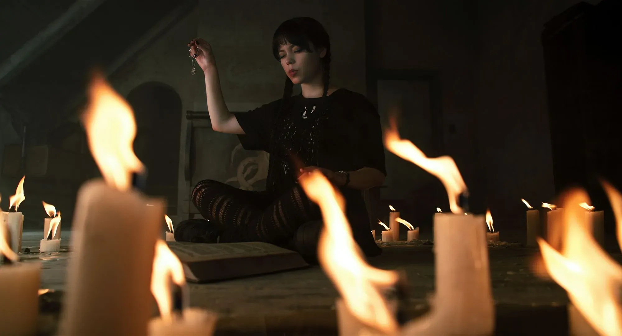 جينا أورتيجا تجلس في غرفة مظلمة محاطة بشموع مضاءة، تظهر بمظهر "الفتاة المتعبة" مع مكياج داكن وعينين مرهقتين، مما يعكس اتجاهات الجمال الجديدة.