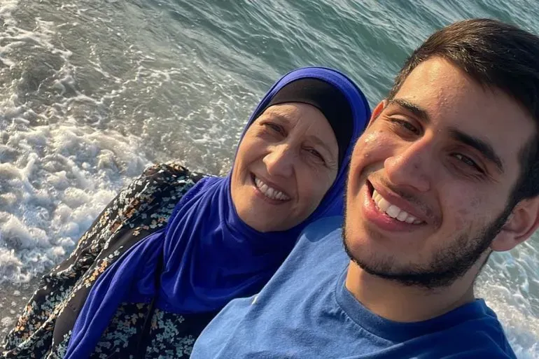 صورة تظهر شابًا مبتسمًا بجانب امرأة مسنّة على شاطئ البحر، مع أمواج تتلاطم خلفهما، تعكس لحظة سعيدة بين العائلة.