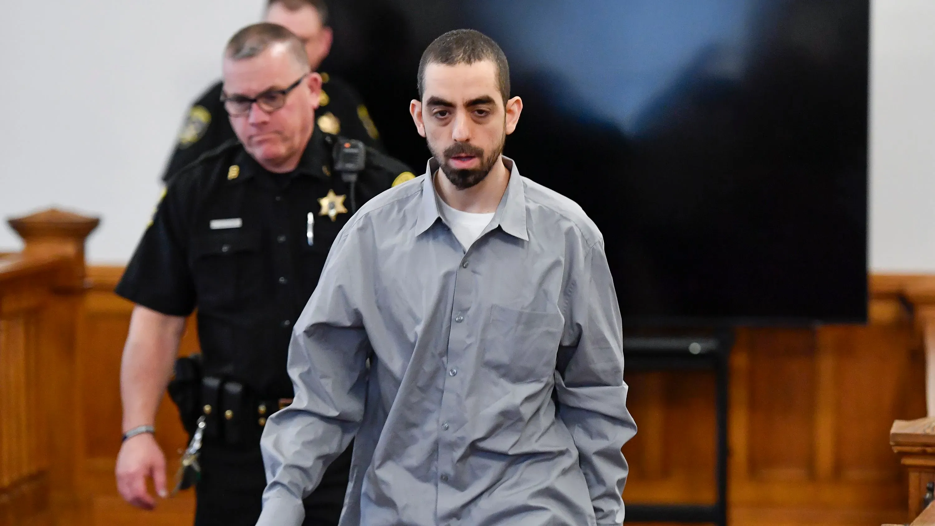 هادي مطر، المدان بالشروع في قتل سلمان رشدي، يدخل قاعة المحكمة برفقة ضباط الشرطة، مع عرض شاشة خلفه.