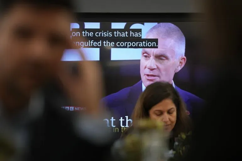 تظهر الصورة شخصًا يتحدث في برنامج إخباري على شاشة تلفزيونية، مع وجود مشاهدين في المقدمة. تتعلق الفضيحة بتأثيرات روبي غيب على تغطية بي بي سي.