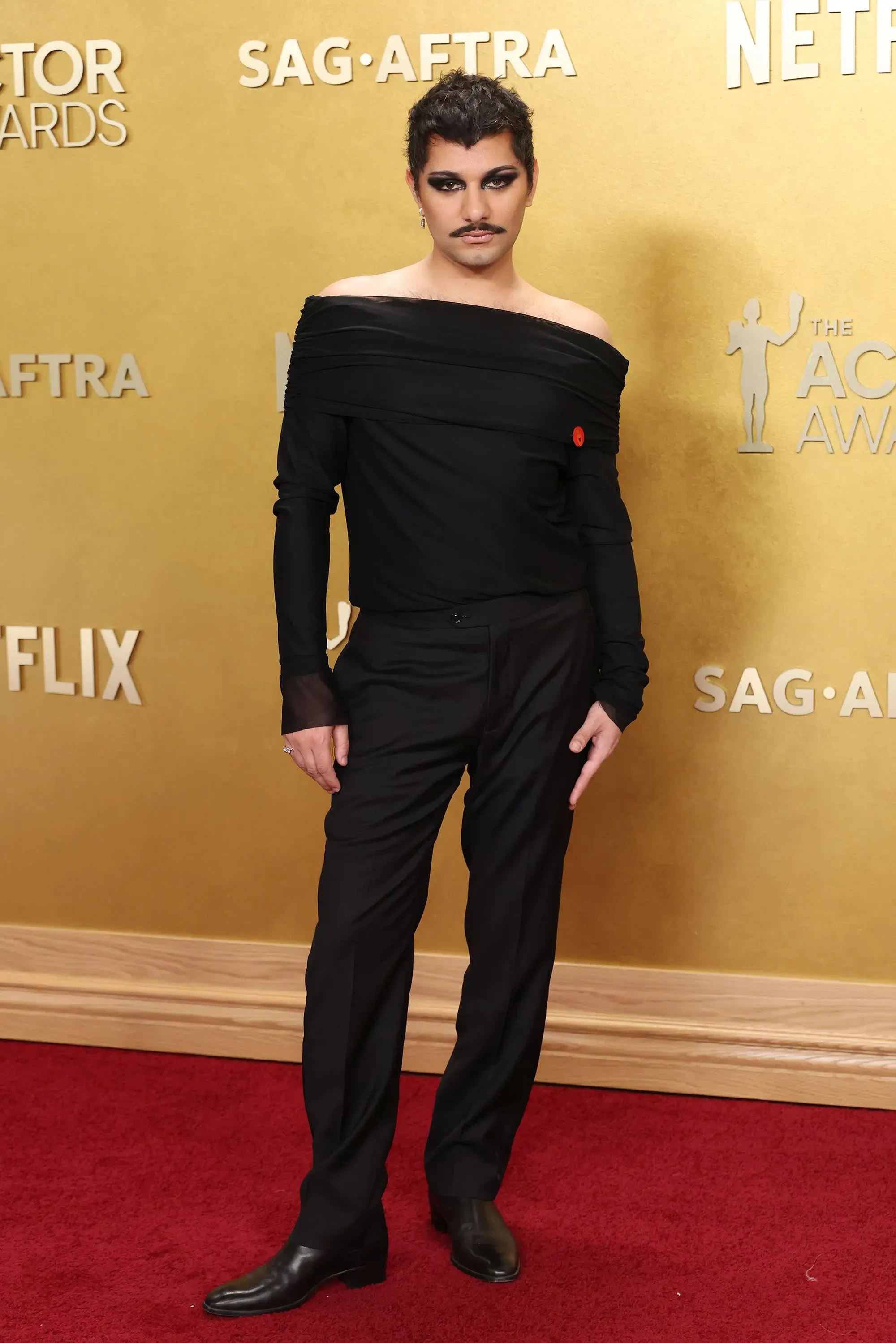 شخص يرتدي ملابس سوداء مبتكرة مع تسريحة شعر مميزة، يقف على السجادة الحمراء في حفل جوائز الممثلين، مع خلفية ذهبية تحمل شعار "نتفليكس".