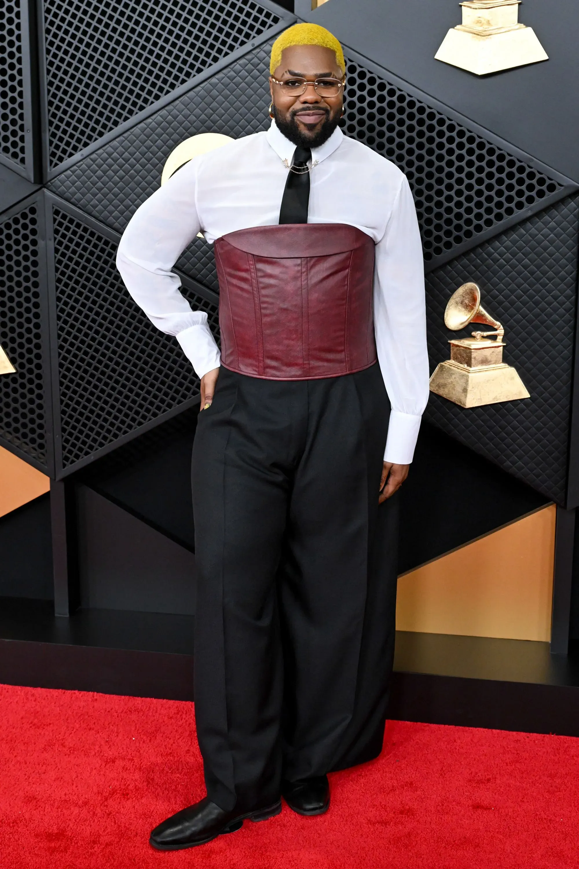 شخص يرتدي سترة كورسيه حمراء وقميص أبيض وبنطلون واسع، يقف على السجادة الحمراء لحفل توزيع جوائز غرامي في لوس أنجلوس.