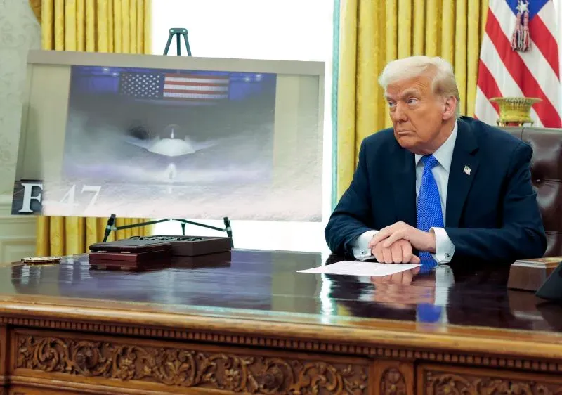 صورة للرئيس ترامب أثناء اجتماع في المكتب البيضاوي، بجانبه صورة لطائرة مقاتلة جديدة تحمل اسم F-47، مع العلم الأمريكي في الخلفية.