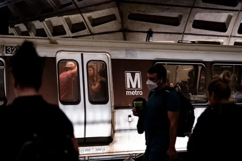 قطار مترو يحمل علامة "M" في محطة، مع ركاب يرتدون أقنعة، يعكس اقتراحات لإعادة تسمية المترو باسم "قطار ترامب".