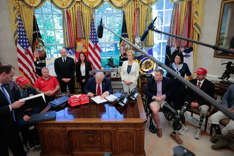 ترامب يوقع وثائق في المكتب البيضاوي محاطًا بمساعدين ومؤيدين، مع ظهور قبعات "اجعل أمريكا عظيمة مرة أخرى" على الطاولة.