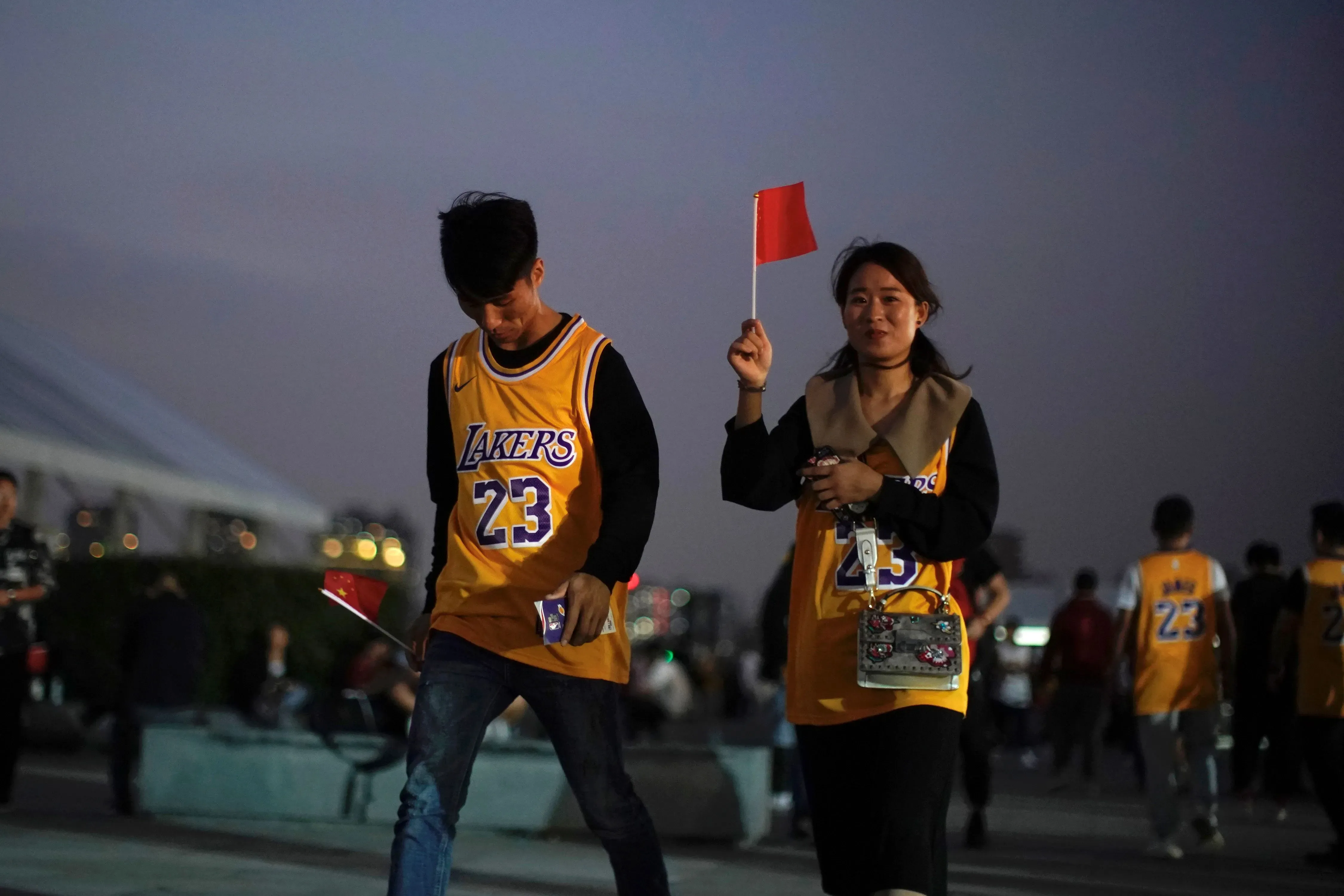شخصان يرتديان قمصان فريق لوس أنجلوس ليكرز، يحمل أحدهما علمًا صغيرًا، في أجواء احتفالية بمناسبة عودة الدوري الأمريكي للمحترفين إلى الصين.