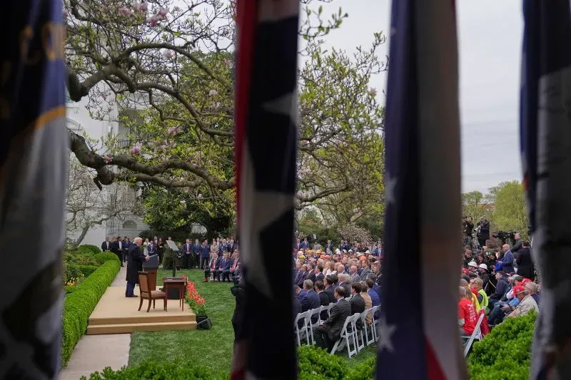 مشهد من حديقة البيت الأبيض خلال حدث رسمي، حيث يتحدث مسؤول أمام جمهور كبير، يحيط به أعلام وطنية وأشجار مزهرة.