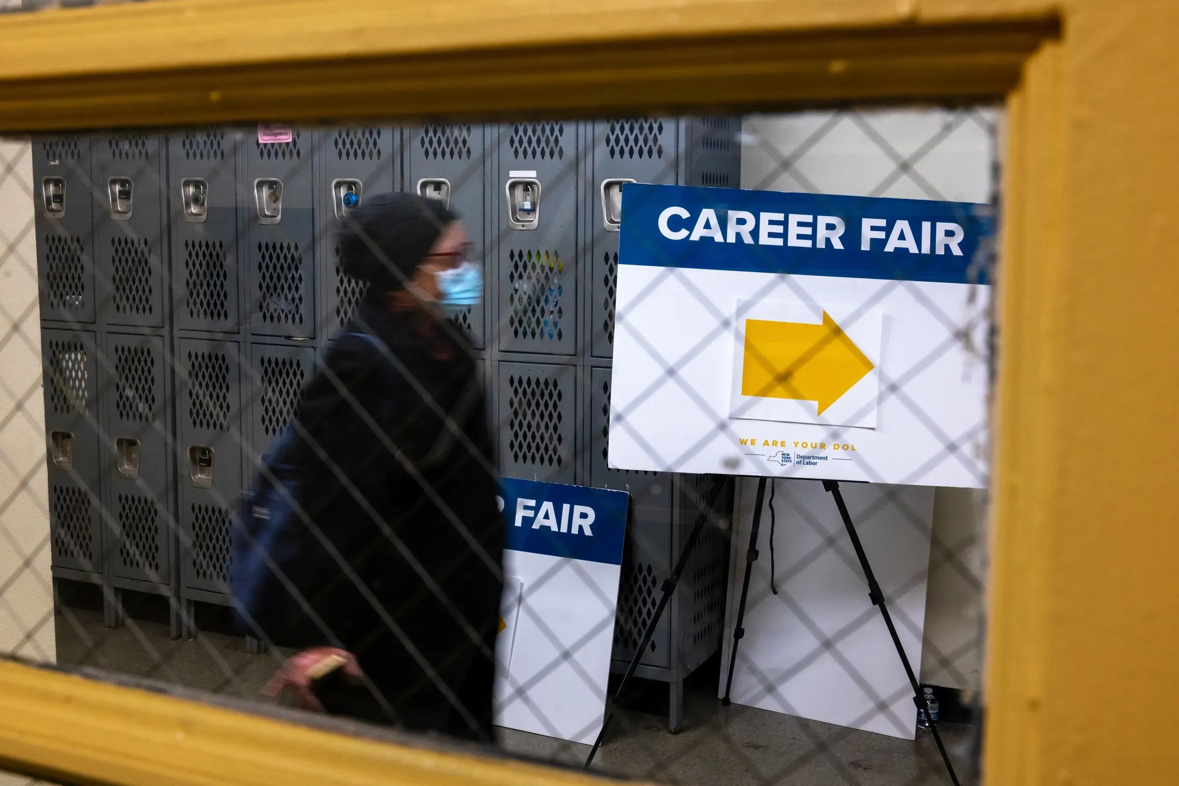 امرأة ترتدي كمامة تسير بجوار لافتة تشير إلى معرض الوظائف، مع وجود خزائن معدنية في الخلفية، مما يعكس تحديات سوق العمل الحالي.