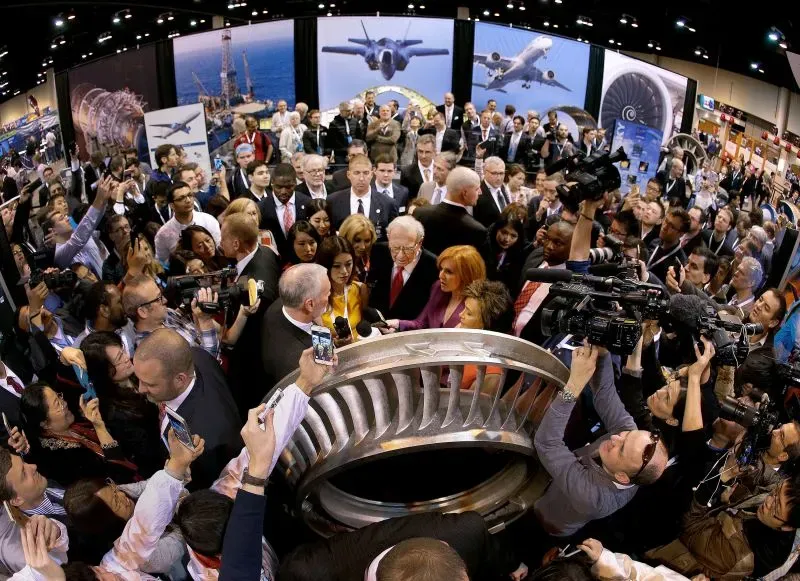 اجتماع حاشد لمساهمي بيركشاير هاثاواي مع وارن بافيت في المركز، محاطاً بالمعجبين ووسائل الإعلام، مع عرض لمعدات الشركات في الخلفية.