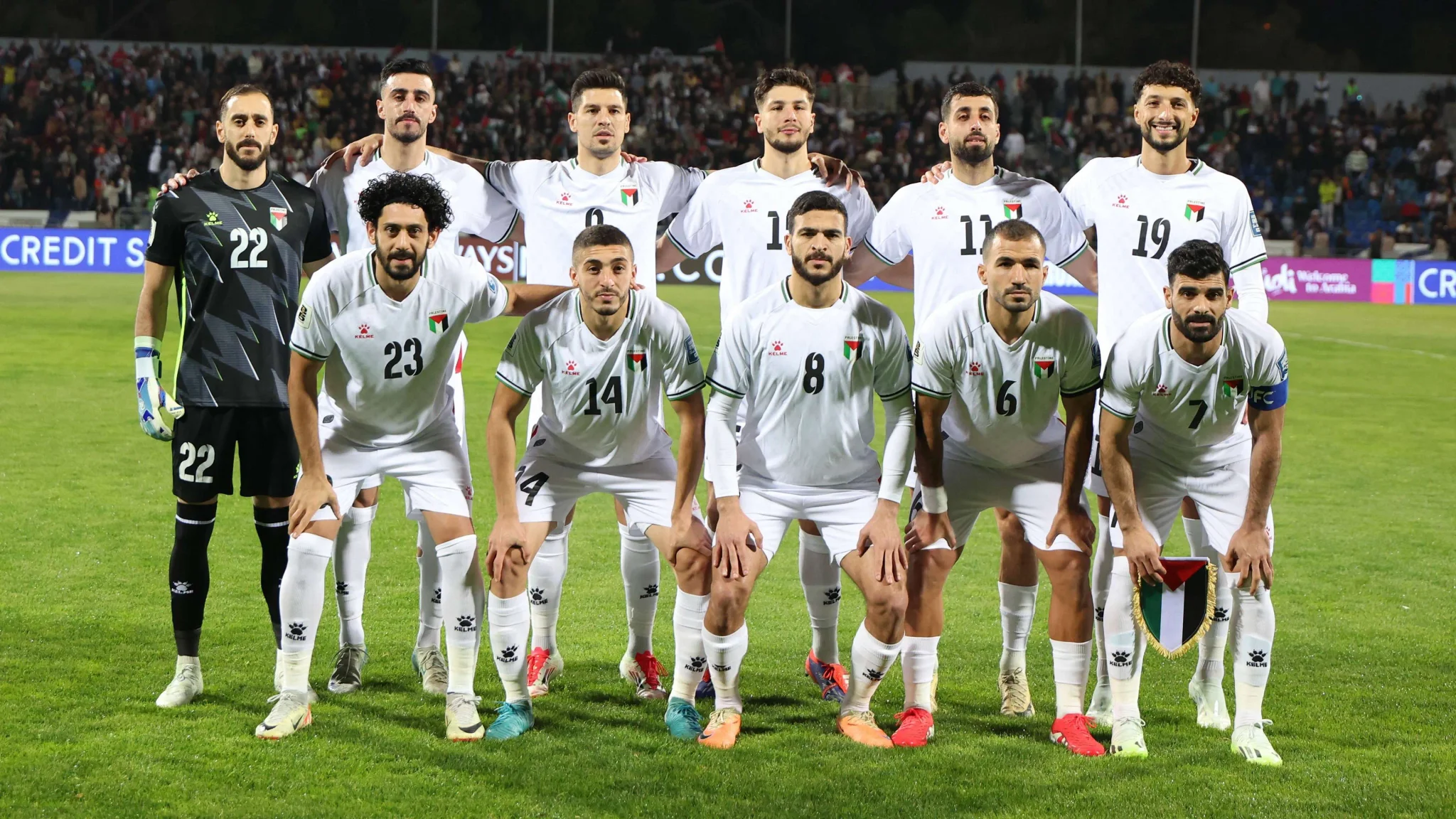 لاعبو المنتخب الفلسطيني لكرة القدم يرتدون الزي الرسمي ويصطفون في ملعب قبل مباراة دولية، مع التركيز على تحديات كرة القدم الفلسطينية ونداءات الإصلاح في FIFA.